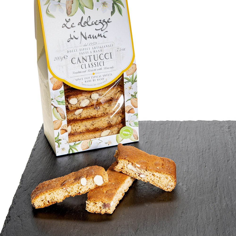 Een vensterverpakking van italiaanse koekjes Cantucci Classici van Le dolcezze di Nanni. Voor de verpakking liggen drie cantucci koekjes. Eentje heeft een hap eruit.